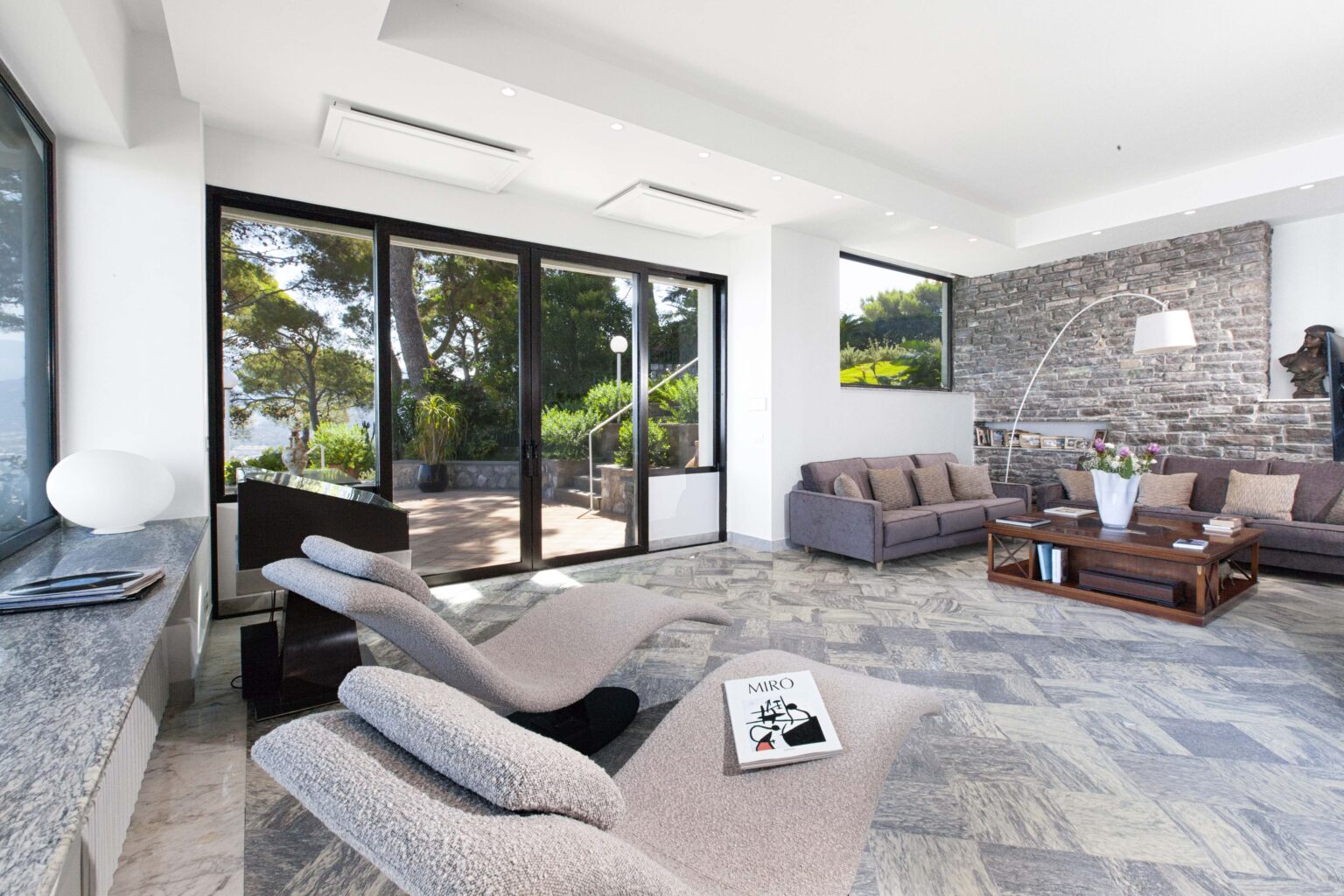 Modern living room featuring large glass doors that open to a scenic outdoor view, stylish gray sofas, a wooden coffee table, and a decorative stone wall. Natural light floods the space, enhancing the contemporary design and inviting atmosphere. | Only on SopranoVillas