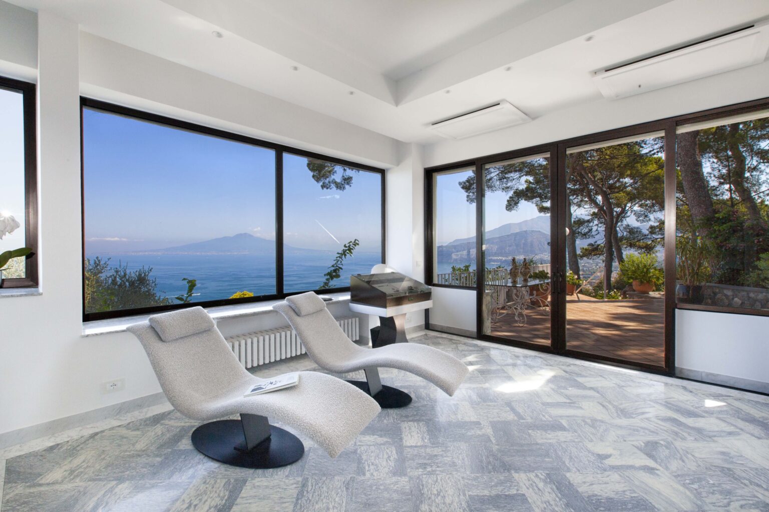 Modern indoor relaxation area with two lounge chairs, large windows showcasing a stunning ocean view, and a serene outdoor terrace surrounded by trees. | Only on SopranoVillas