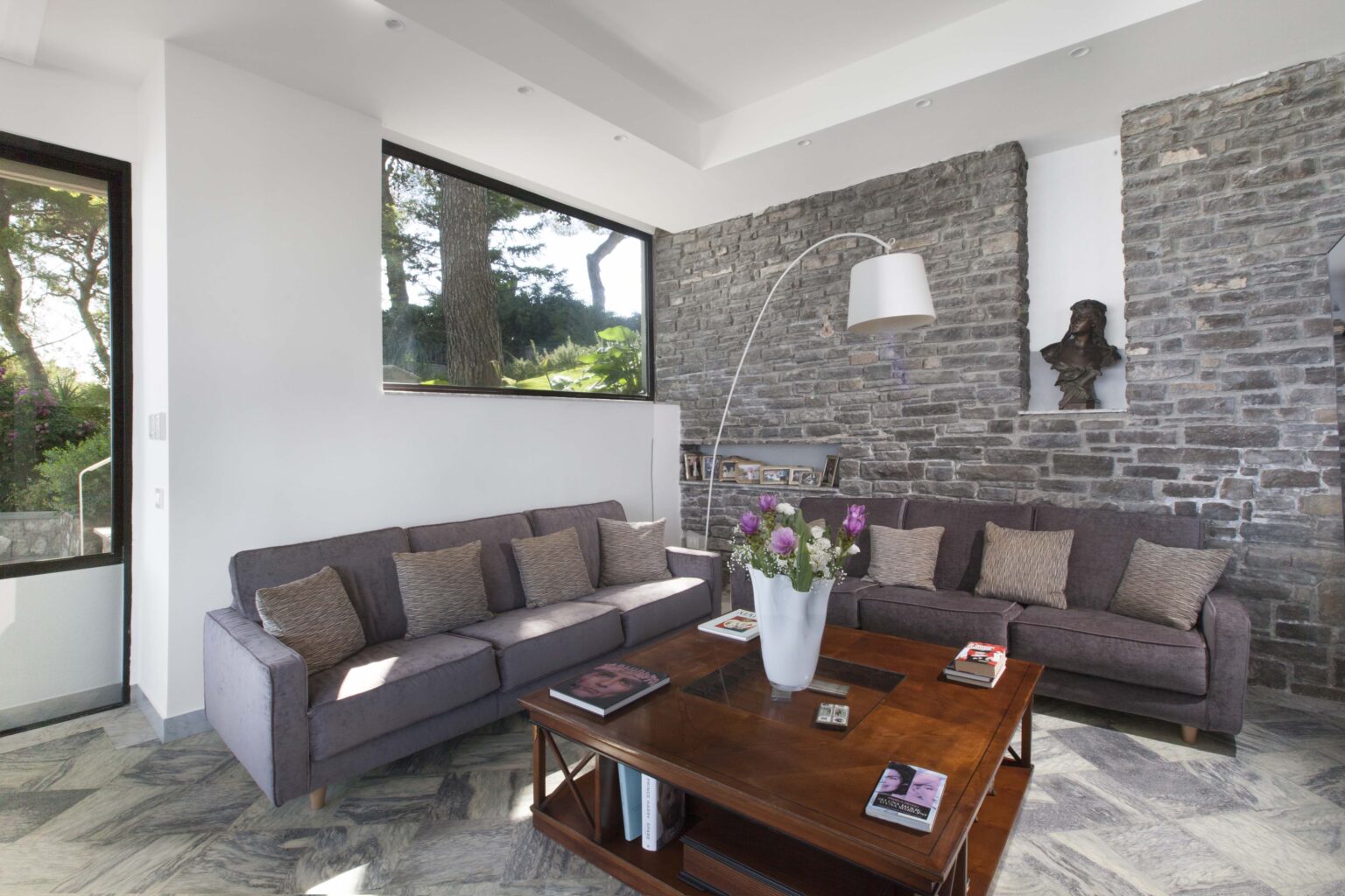 Modern living room featuring a gray sectional sofa, a wooden coffee table with magazines, and a stylish floor lamp. Large windows provide natural light and a view of greenery, enhancing the contemporary design with stone wall accents. | Only on SopranoVillas