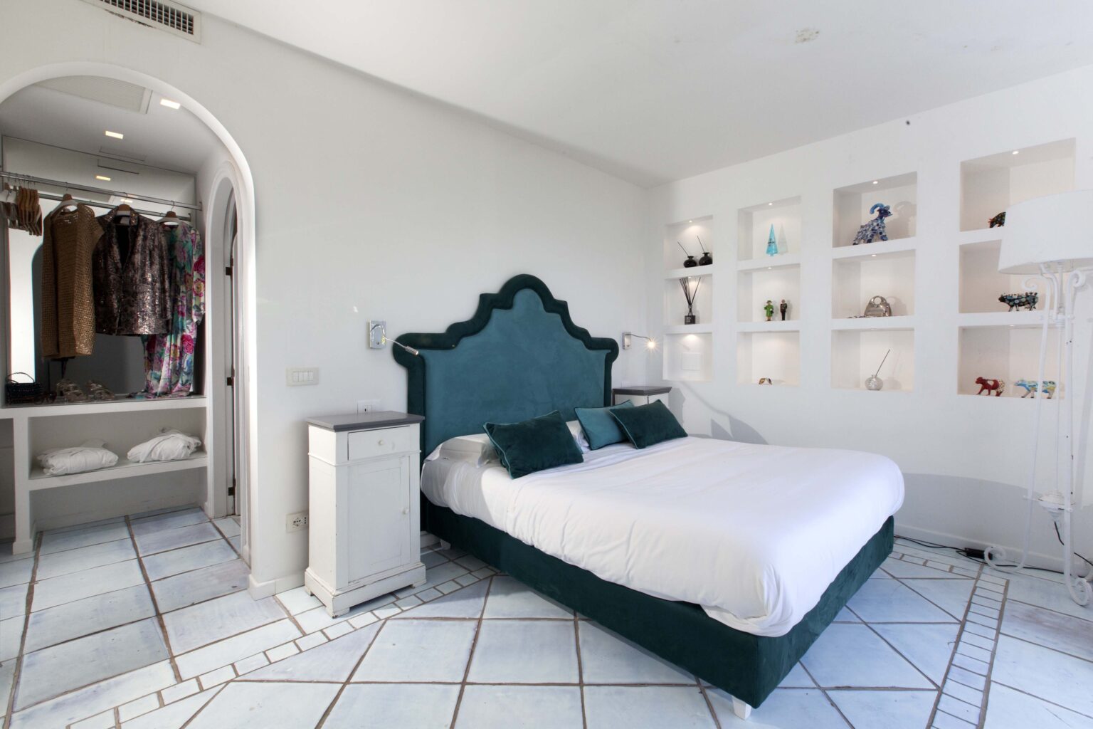 Modern bedroom featuring a teal upholstered bed, white bedding, and stylish decor. The room includes a walk-in closet, open shelving with decorative items, and a tiled floor, creating a bright and inviting atmosphere. | Only on SopranoVillas