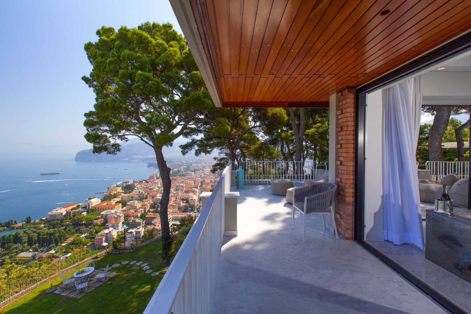 Modern balcony view overlooking a coastal town and the sea, surrounded by lush trees and bright blue skies. The stylish outdoor seating area features contemporary furniture, creating an inviting space for relaxation. | Only on SopranoVillas