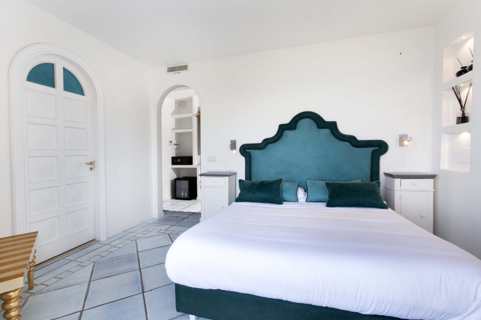 Modern bedroom with a stylish green headboard, white bedding, and minimalist decor. Features a light-filled space with arched doorways and a small kitchenette in the background. Ideal for a relaxing getaway. | Only on SopranoVillas