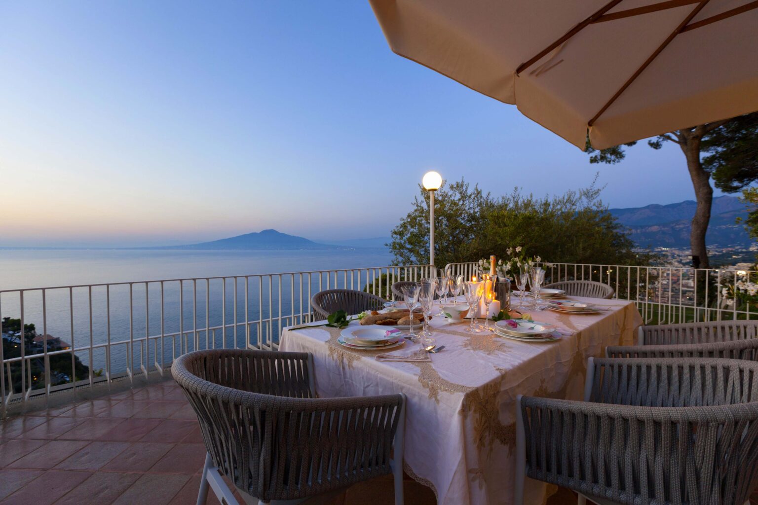 Elegant outdoor dining setup overlooking the ocean at sunset, featuring a beautifully arranged table with fine dining elements and a view of a distant volcano. | Only on SopranoVillas