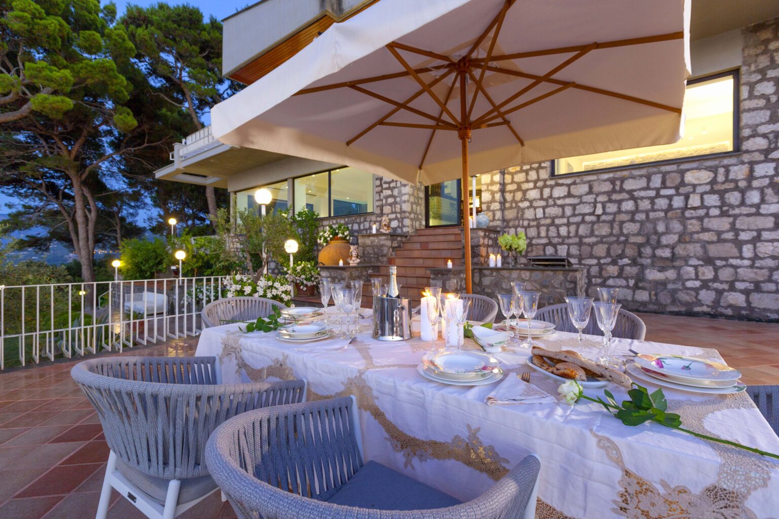 Elegant outdoor dining setup featuring a beautifully arranged table with fine china, crystal glassware, and a centerpiece of fresh flowers, set against a backdrop of a modern stone house and lush greenery. The scene is illuminated by soft lighting, creating a cozy atmosphere perfect for evening meals. | Only on SopranoVillas