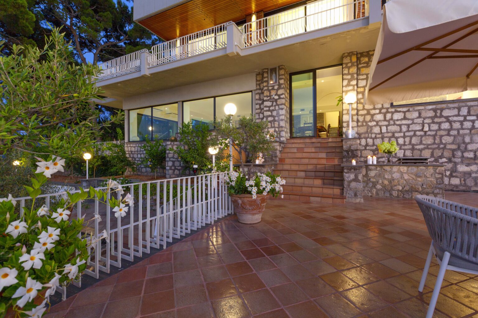 Modern stone house exterior with a spacious patio, lush greenery, and decorative flowers, illuminated at dusk. The image showcases steps leading to large glass windows, enhancing the inviting atmosphere of the property. | Only on SopranoVillas