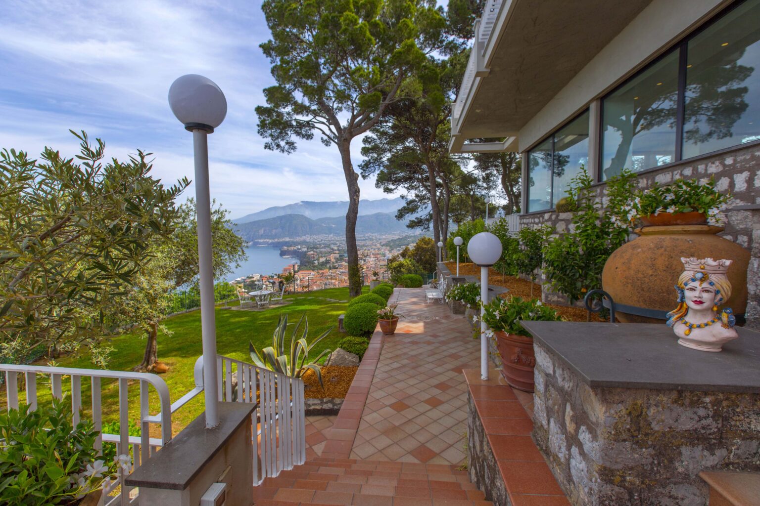 Scenic view from a hillside terrace featuring well-maintained gardens, stone pathways, and decorative planters, overlooking a lake and mountains under a clear sky. | Only on SopranoVillas
