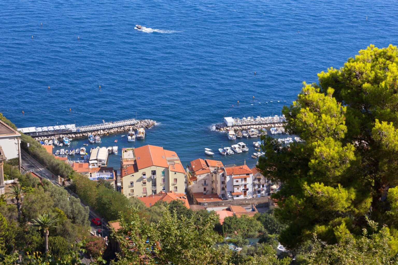 Aerial view of a coastal town with colorful buildings and a marina, situated near a blue sea, featuring boats and lush greenery in the foreground. | Only on SopranoVillas