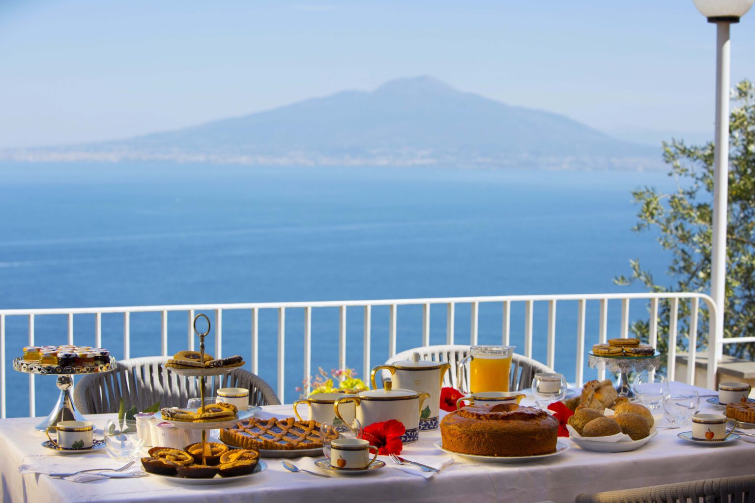 A picturesque outdoor dining setup featuring a variety of Italian pastries and desserts, with a stunning view of Mount Vesuvius and the sea in the background. The table is elegantly arranged with plates of sweets, a glass of juice, and decorative flowers, creating a serene and inviting atmosphere. | Only on SopranoVillas