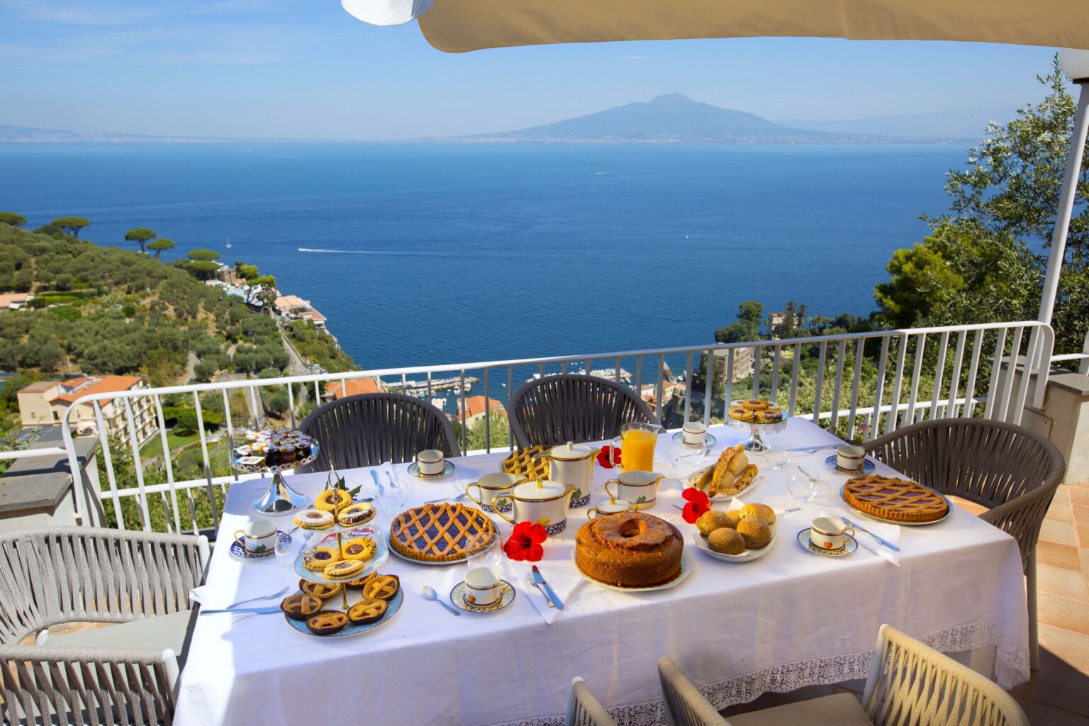 Elegant outdoor breakfast setting with a variety of pastries and drinks on a table, overlooking a scenic ocean view and a distant volcano. Ideal for showcasing luxury dining experiences in coastal locations. | Only on SopranoVillas