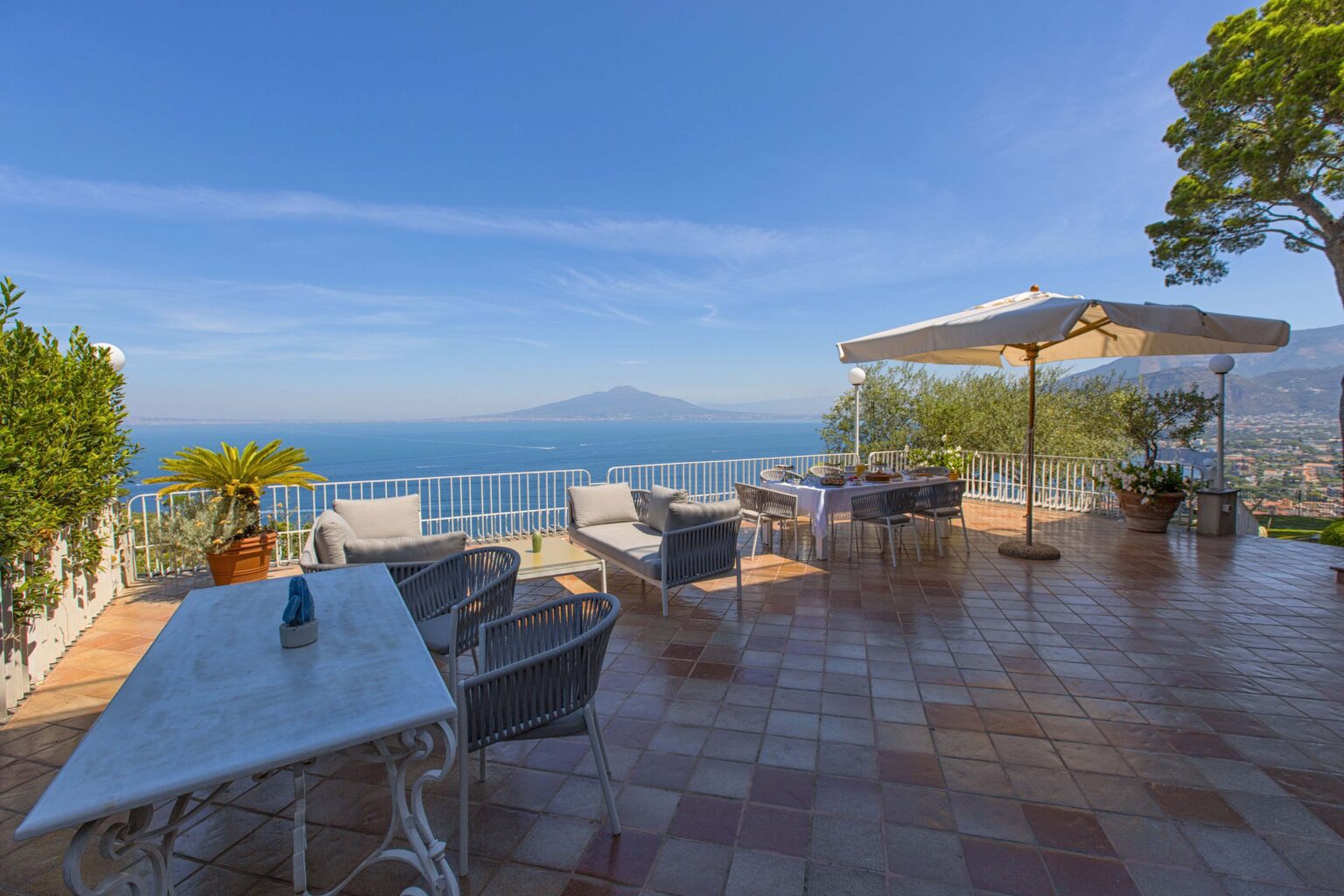 Scenic terrace with outdoor seating overlooking the ocean and mountains, featuring a large umbrella for shade and vibrant greenery. Ideal for relaxation and enjoying coastal views. | Only on SopranoVillas