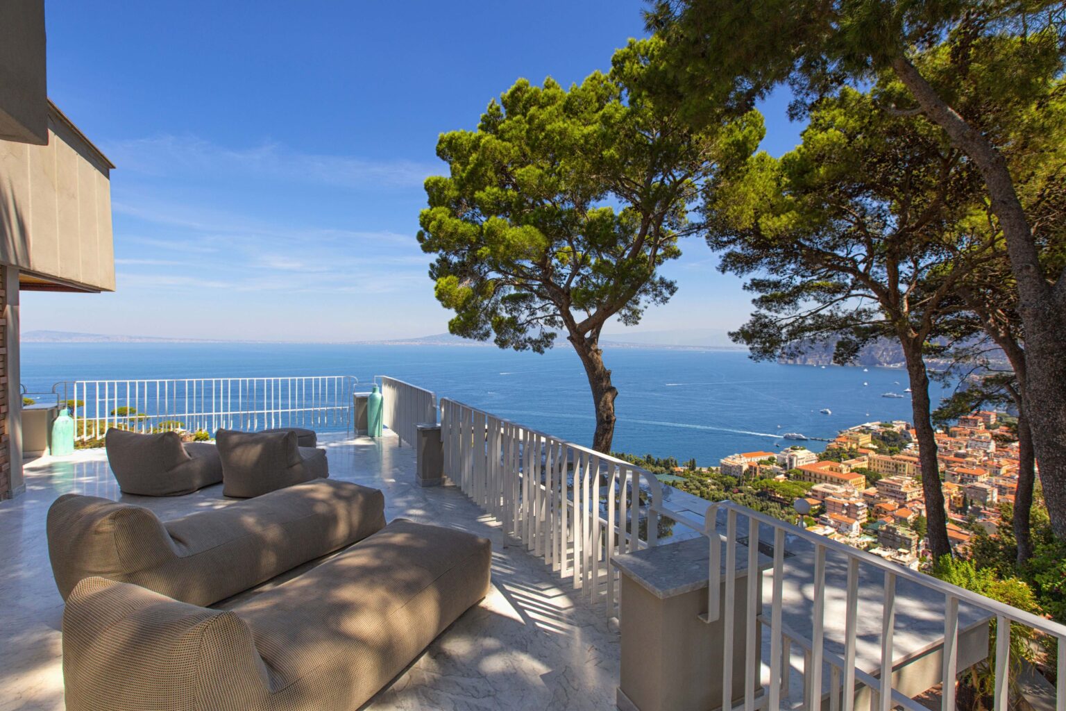 Scenic terrace with modern seating overlooking the ocean, surrounded by lush pine trees and a clear blue sky. Ideal for relaxation and enjoying coastal views. | Only on SopranoVillas