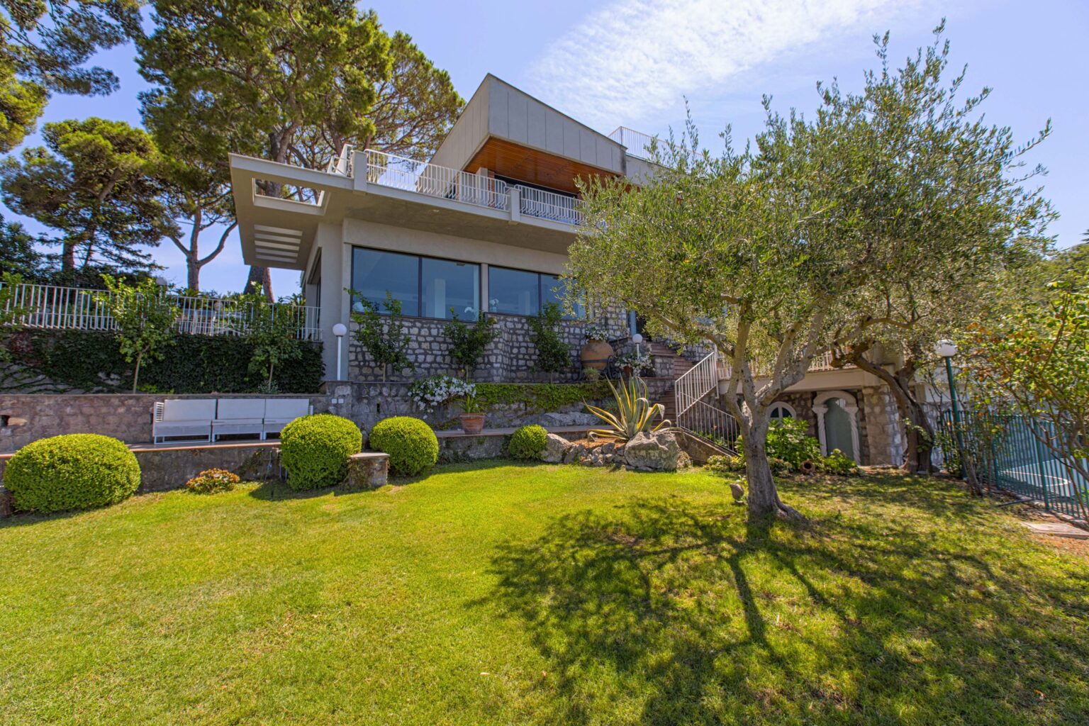 Modern villa with large windows surrounded by lush greenery and landscaped garden, featuring olive trees and well-maintained shrubs under a clear blue sky. | Only on SopranoVillas