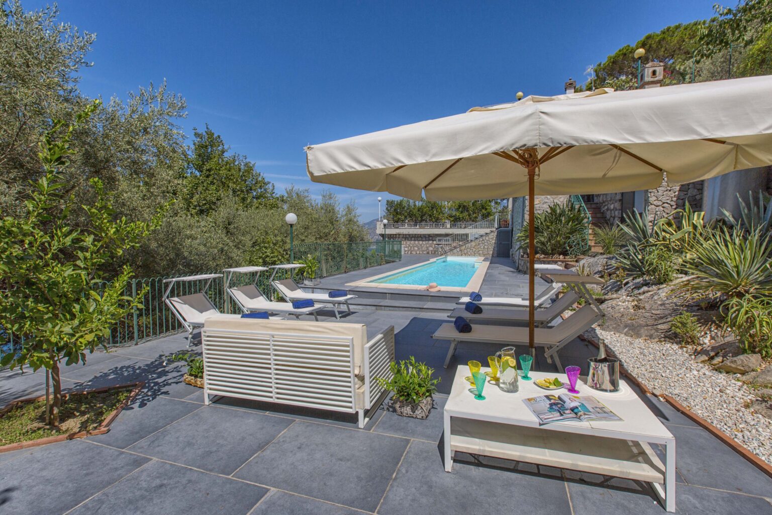 Luxurious outdoor patio with a swimming pool, sun loungers, and an umbrella, surrounded by greenery and stone pathways, perfect for relaxation and leisure. | Only on SopranoVillas