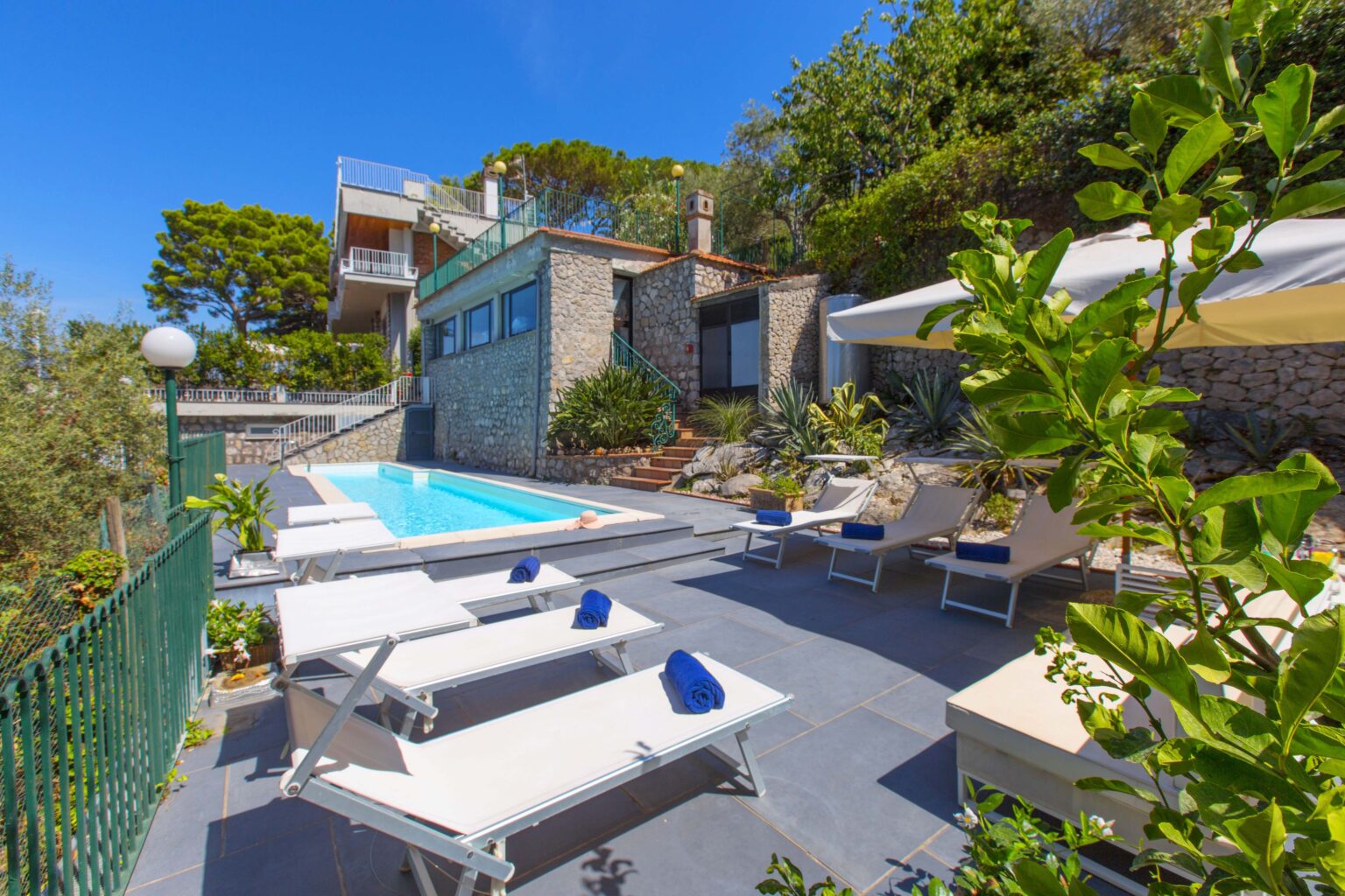 Luxurious outdoor pool area featuring comfortable lounge chairs with blue towels, surrounded by lush greenery and a modern stone villa in a sunny setting. Ideal for relaxation and leisure activities. | Only on SopranoVillas