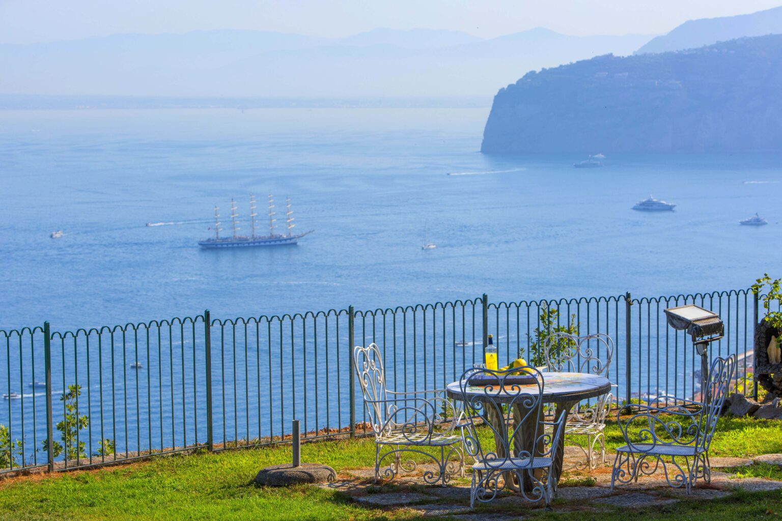 Scenic view of a coastal landscape featuring a table set for two with drinks, overlooking a tranquil sea and distant cliffs, ideal for outdoor dining and relaxation. | Only on SopranoVillas