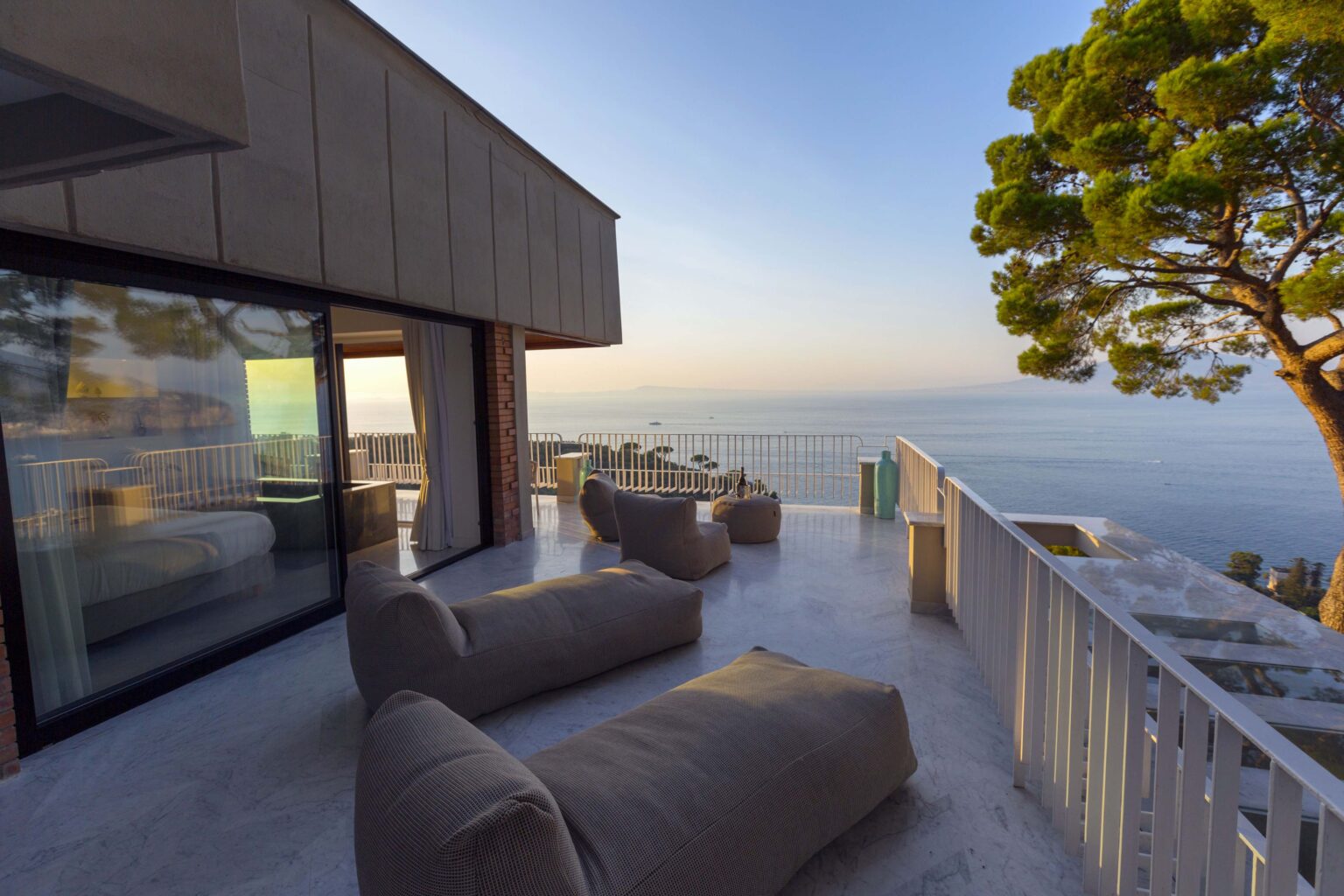 Modern outdoor terrace with comfortable seating, offering a stunning ocean view at sunrise. The space features large glass doors, a minimalist design, and lush greenery, creating a serene atmosphere for relaxation. | Only on SopranoVillas
