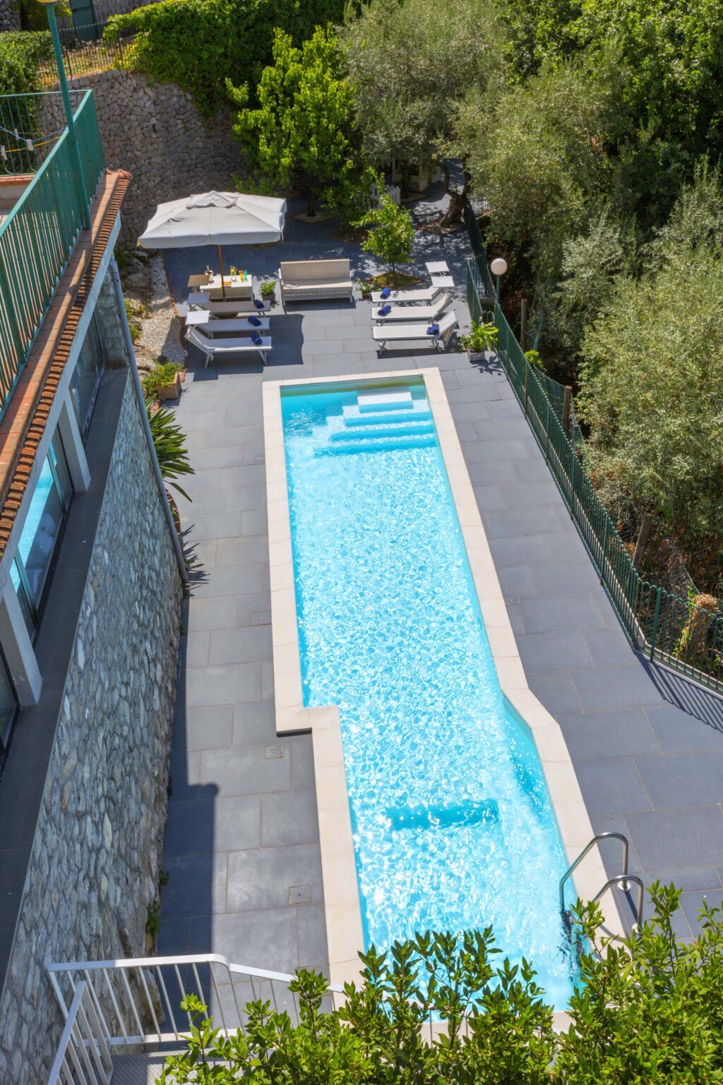 Modern outdoor swimming pool with clear blue water, surrounded by sleek gray tiles and lounge chairs, set in a lush garden environment. Ideal for luxury home design inspiration. | Only on SopranoVillas