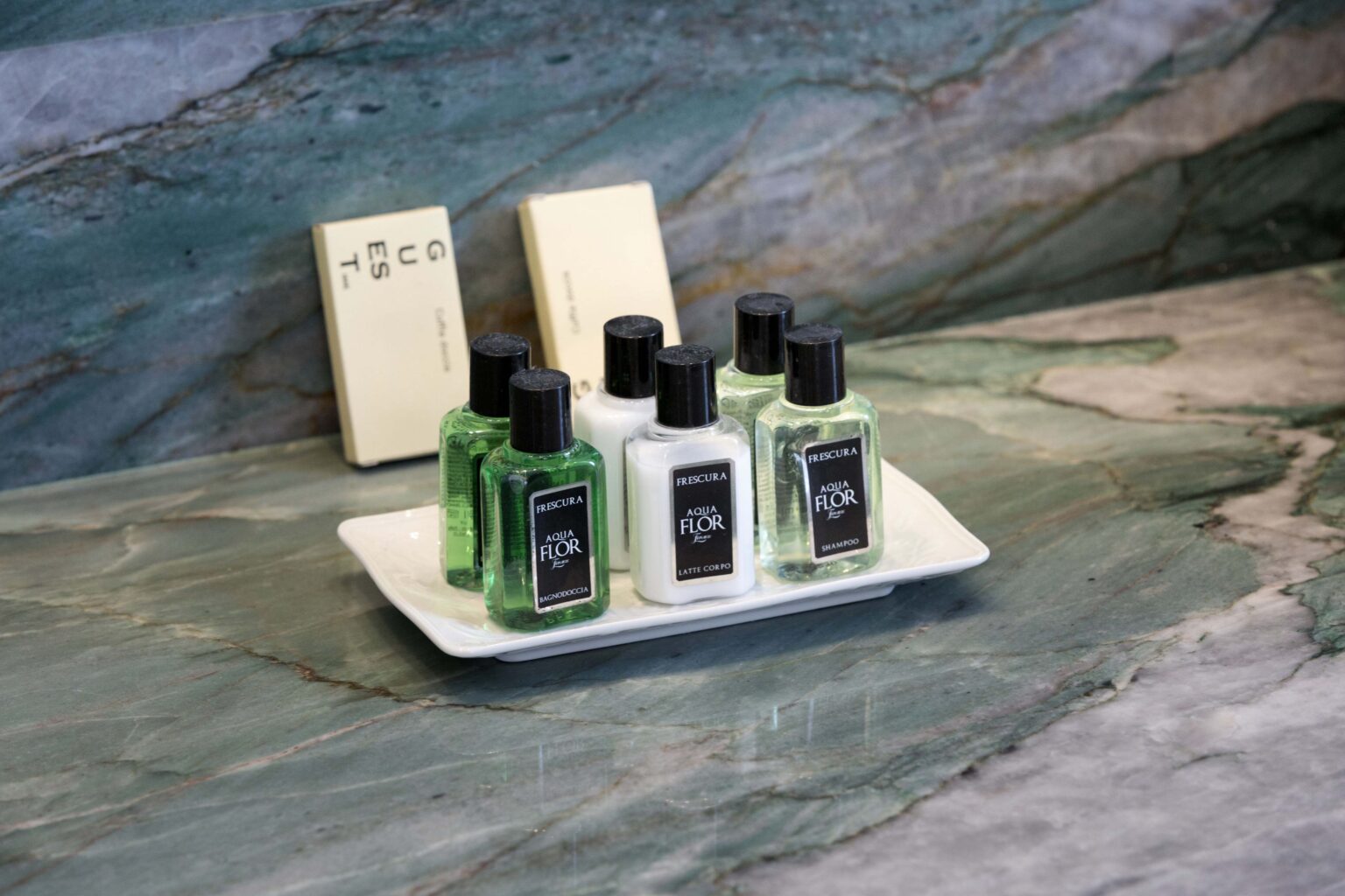 Set of travel-sized hair and body care products displayed on a white tray, featuring green and clear bottles labeled with "Aqua Flor" and "Frescura," placed on a textured green marble countertop alongside two soap bars. | Only on SopranoVillas