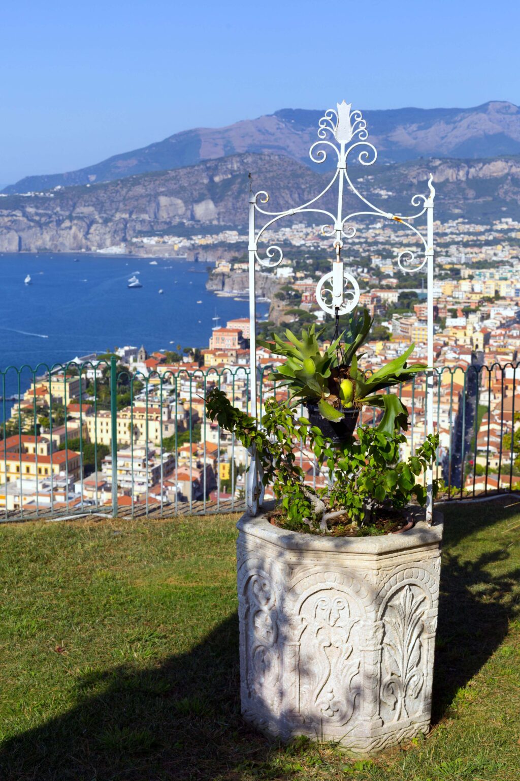 Scenic view of a decorative stone planter with lush greenery, overlooking a coastal town and mountains in the background under a clear blue sky. | Only on SopranoVillas