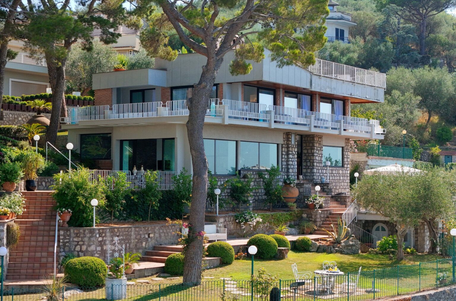 Modern villa with a spacious terrace surrounded by lush greenery and landscaped gardens, featuring stone steps and outdoor seating areas. Ideal for a relaxing getaway. | Only on SopranoVillas