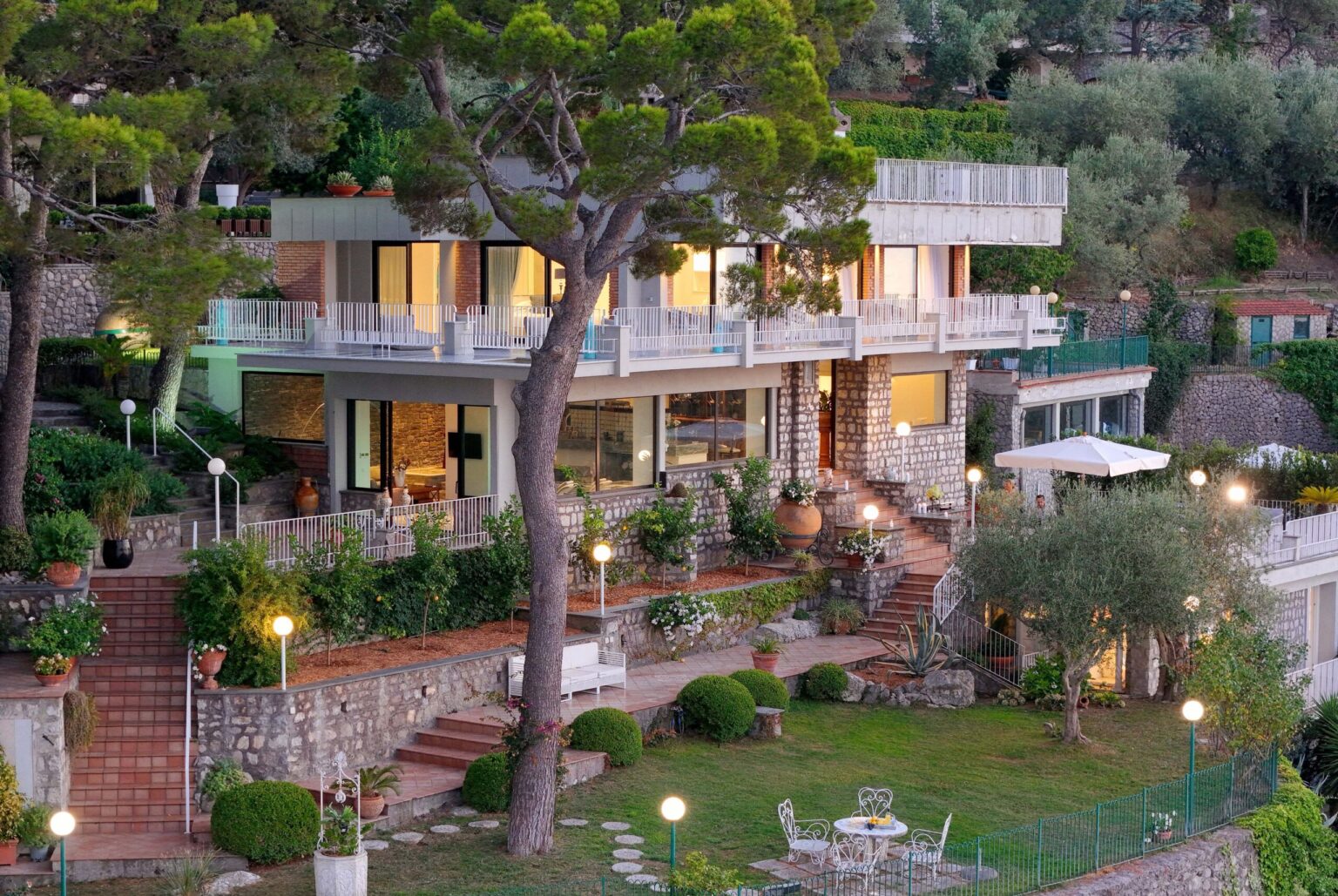 Modern villa surrounded by lush greenery, featuring a well-designed garden, outdoor seating areas, and elegant architecture, illuminated in the evening light. | Only on SopranoVillas