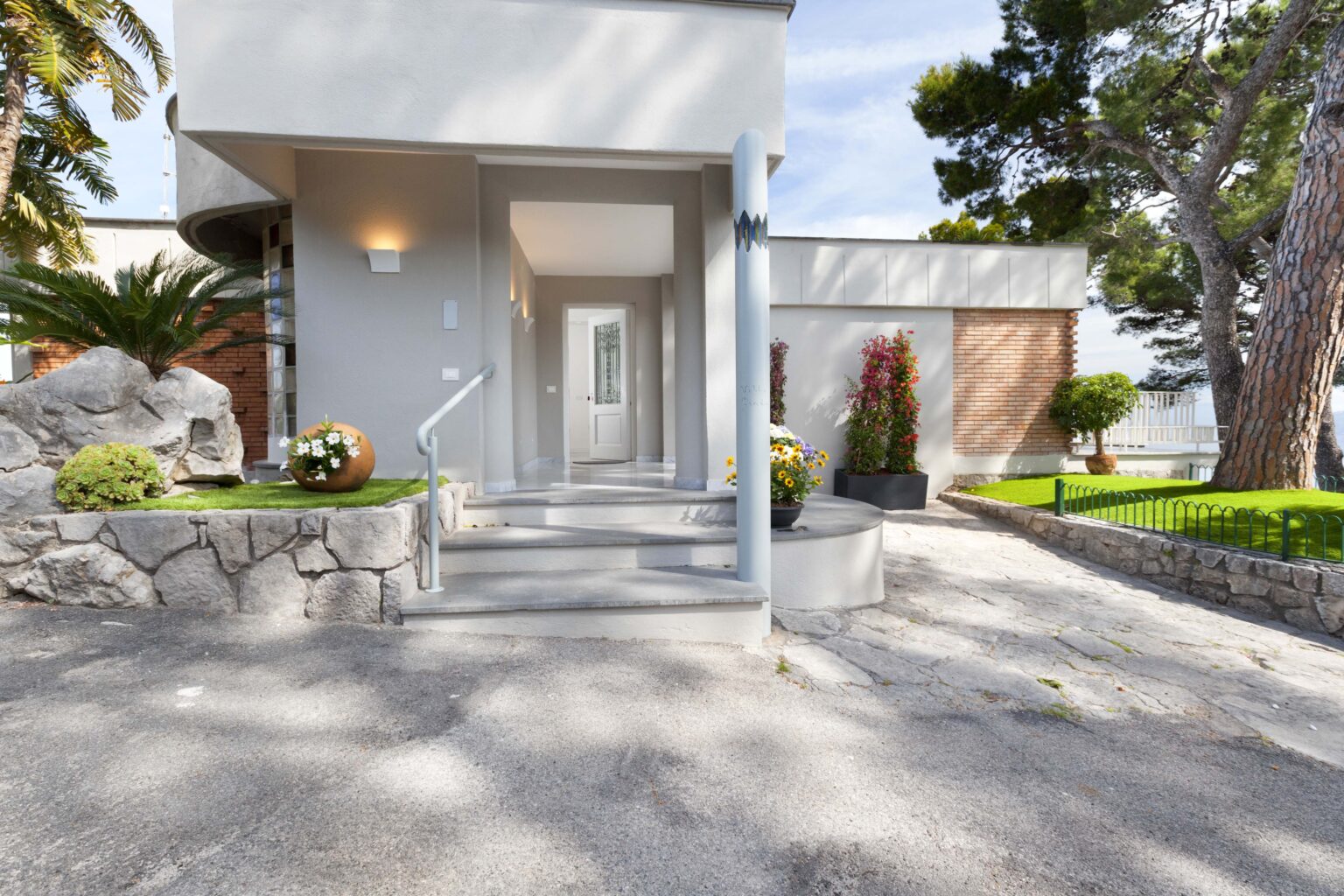 Modern entrance to a contemporary home featuring a landscaped pathway, stone accents, and lush greenery, set in a serene outdoor environment. | Only on SopranoVillas