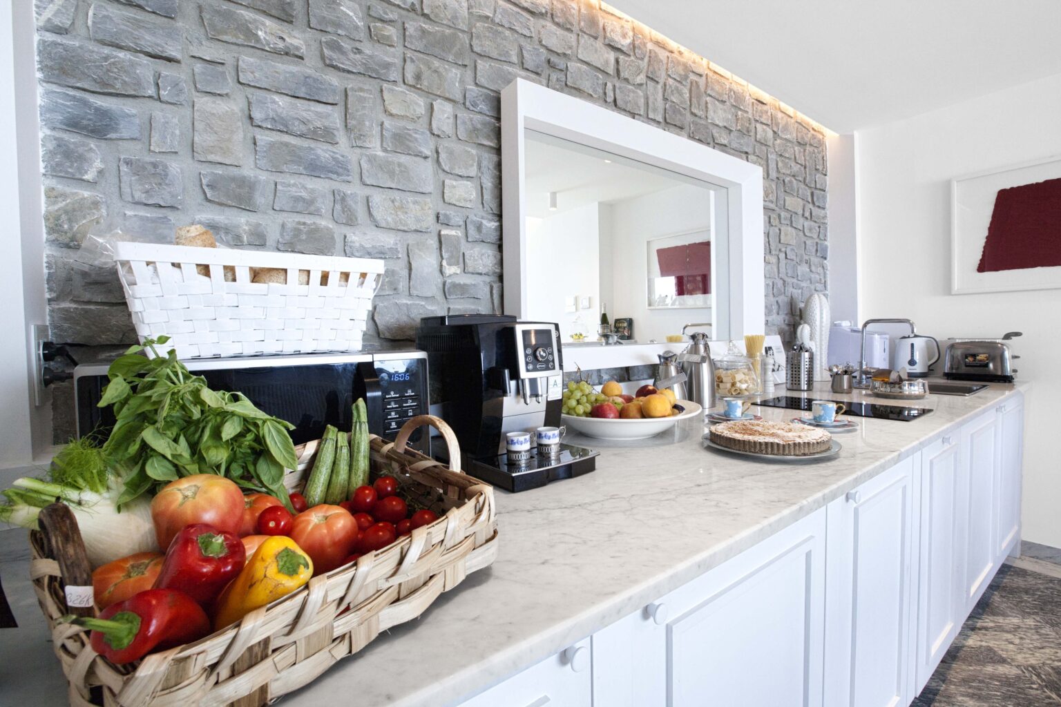 Modern kitchen countertop featuring a stone wall backdrop, a basket of fresh fruits and vegetables, kitchen appliances, and a stylish decor. | Only on SopranoVillas