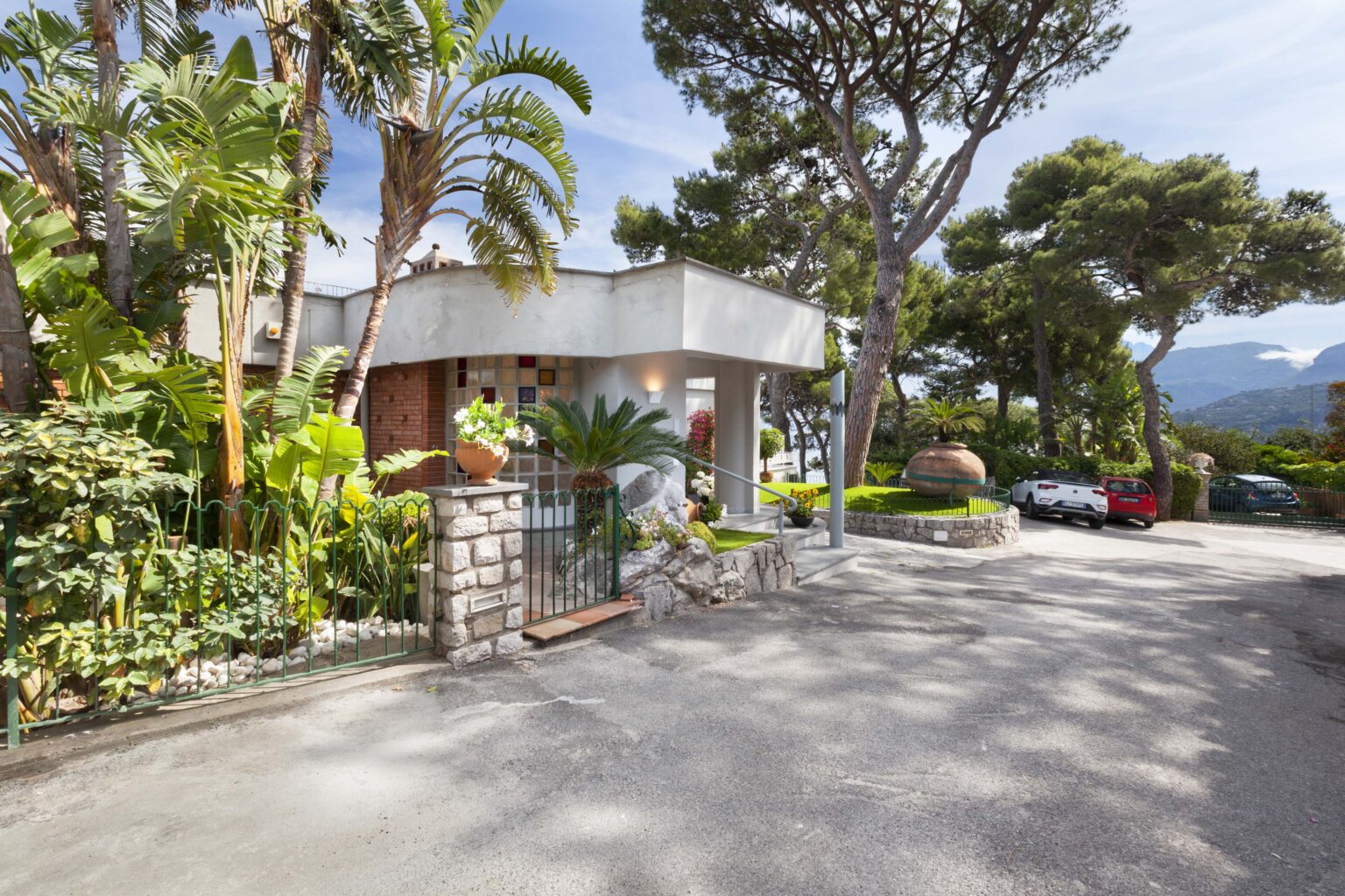 Modern home entrance surrounded by tropical plants, featuring a stone pathway and a driveway with parked cars, set against a backdrop of mountains and blue sky. | Only on SopranoVillas