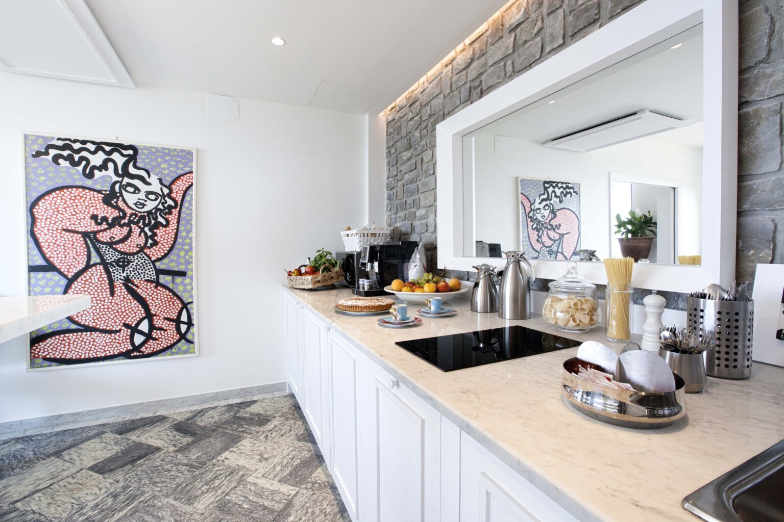 Modern kitchen interior featuring a stone accent wall, sleek white cabinetry, and a large mirror. The countertop displays a variety of fresh fruits and kitchen appliances, with a colorful abstract artwork hanging on the wall. | Only on SopranoVillas