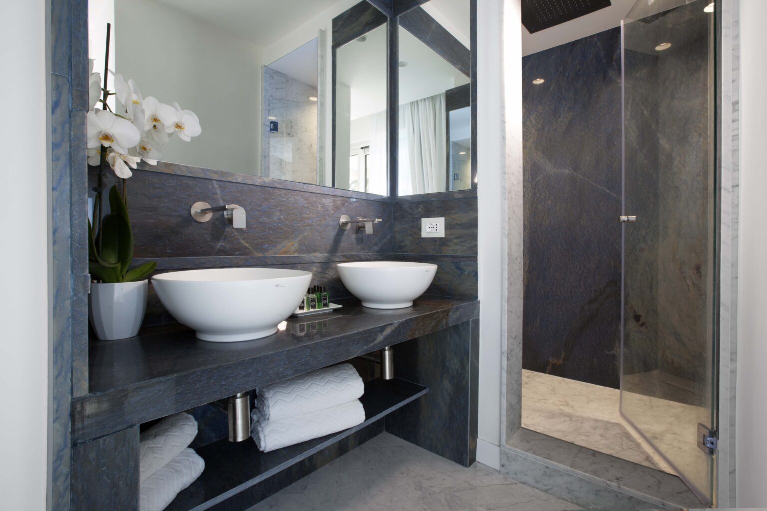 Modern bathroom design featuring two round sinks on a sleek countertop, a glass shower enclosure, and neatly stacked towels, accented by a decorative plant. | Only on SopranoVillas