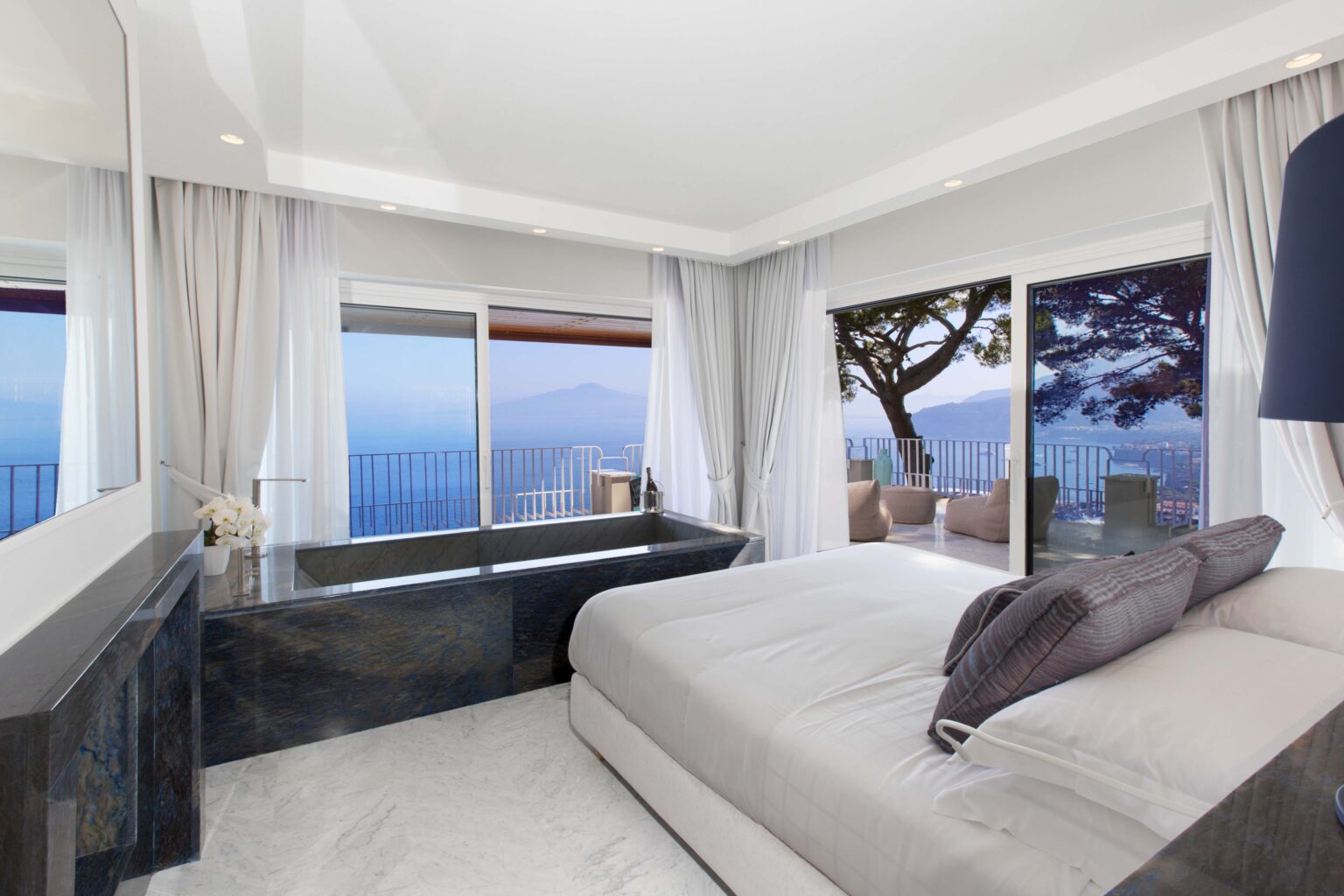 Modern bedroom with a king-sized bed, large windows offering a scenic ocean view, and a stylish black marble bathtub. The room features soft gray tones and elegant curtains, creating a serene and luxurious atmosphere. | Only on SopranoVillas