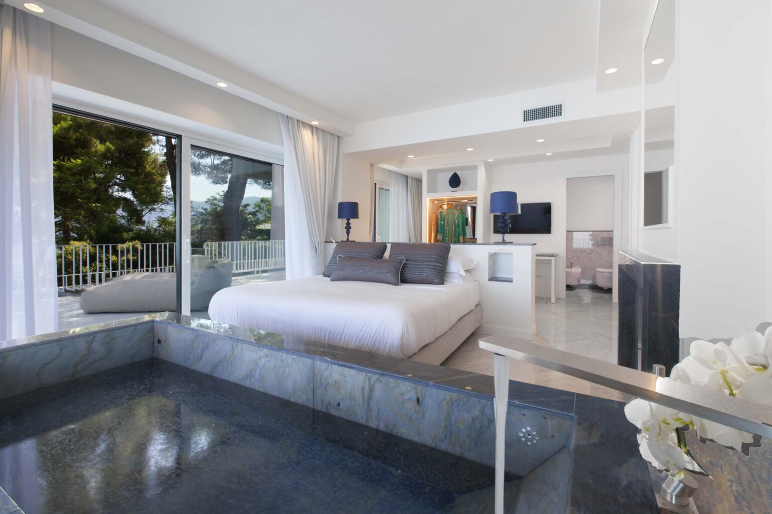 Modern bedroom with a king-sized bed, large windows offering natural light, and an elegant private pool area. The room features minimalist decor with neutral tones, a stylish desk, and a flat-screen TV, creating a serene and luxurious atmosphere. | Only on SopranoVillas