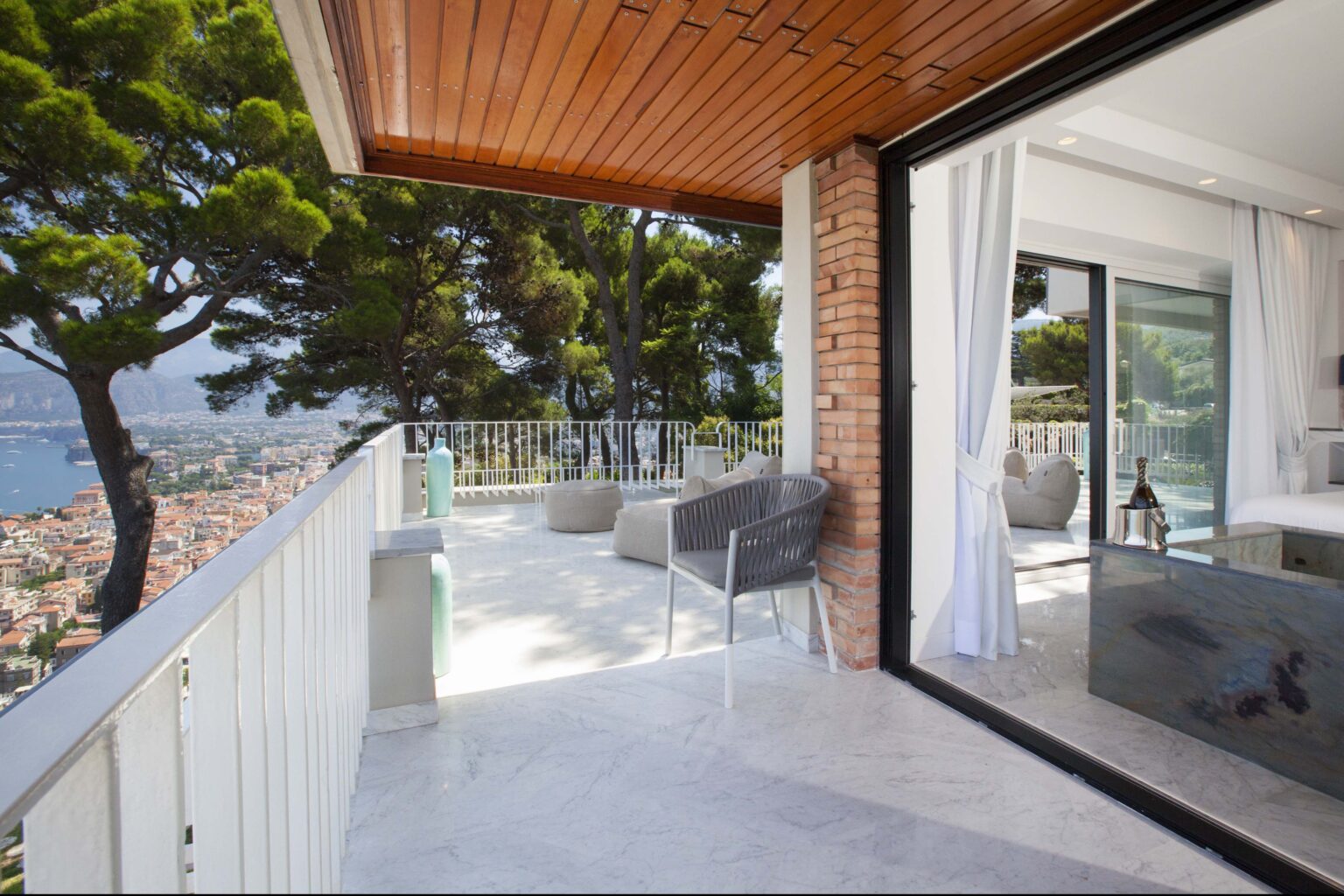 Modern balcony with a scenic view overlooking the city and sea, featuring stylish outdoor seating and a sleek glass door leading to an interior space. | Only on SopranoVillas