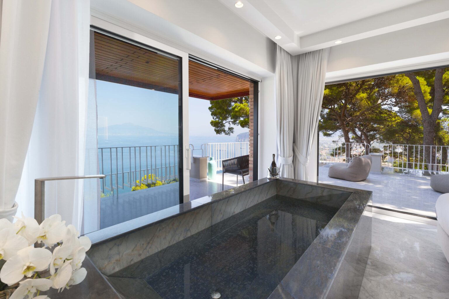 Luxurious bathroom featuring a sleek black stone bathtub with a stunning view of the ocean through large glass windows, surrounded by elegant decor and natural light. | Only on SopranoVillas