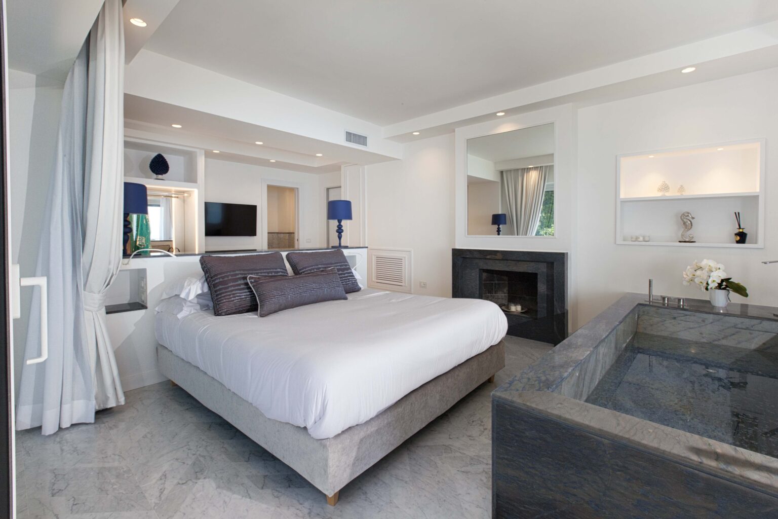 Modern bedroom design featuring a king-size bed with gray linens, minimalist decor, and a cozy seating area. The room includes a fireplace, large windows for natural light, and a stylish bathtub, creating a serene and inviting atmosphere. | Only on SopranoVillas