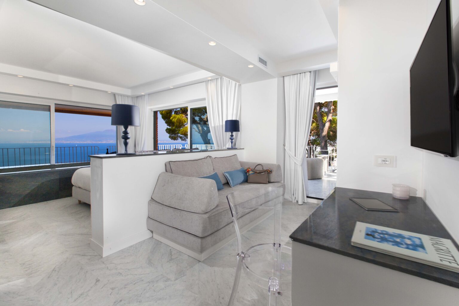 Modern hotel suite with a comfortable gray sofa, elegant decor, and large windows offering a stunning ocean view. Features include a sleek television, minimalist furnishings, and access to an outdoor patio. | Only on SopranoVillas