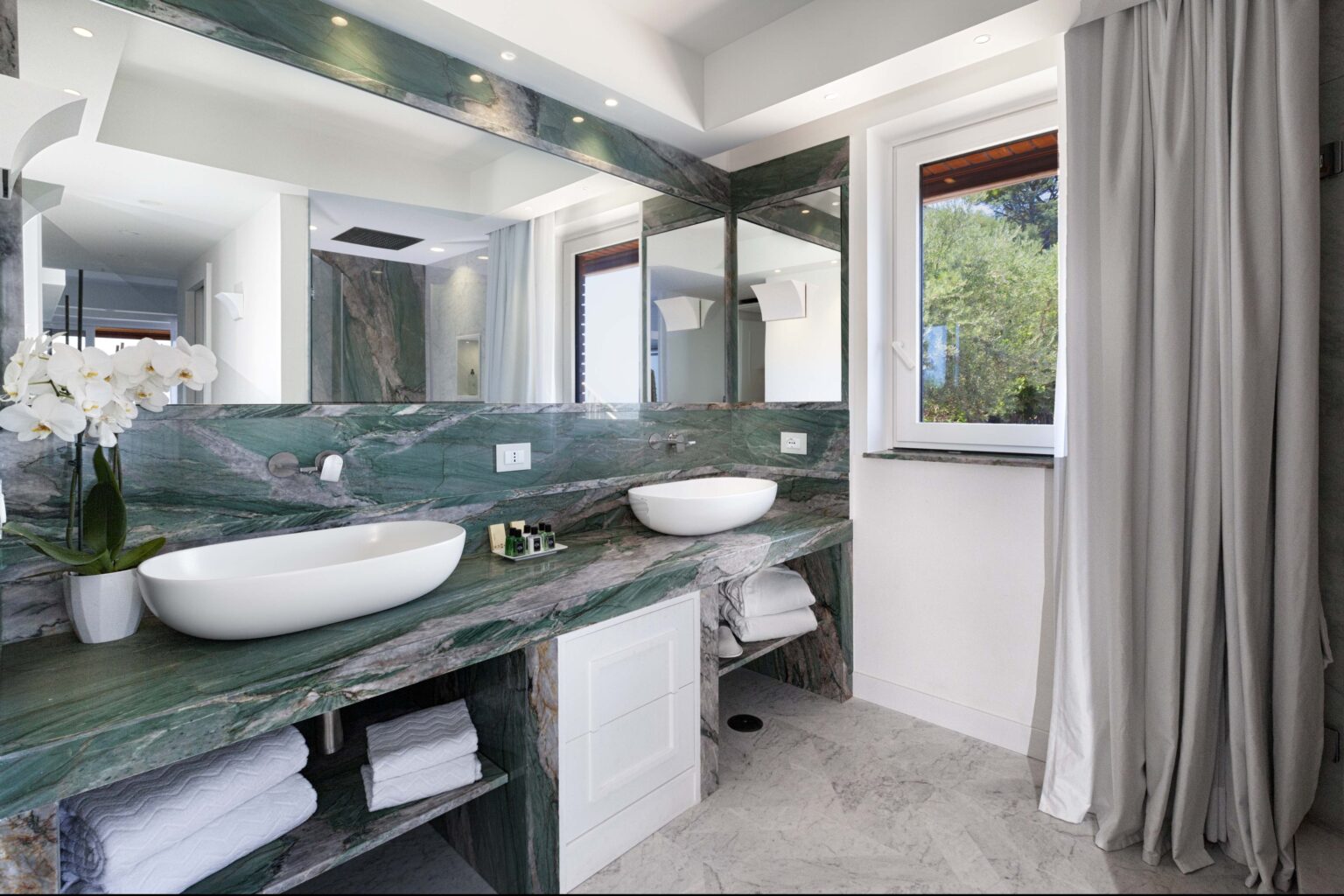 Modern bathroom interior featuring green marble countertops, double sinks, and large mirrors. Natural light floods the space through a window, complemented by elegant decor and neatly arranged towels. | Only on SopranoVillas