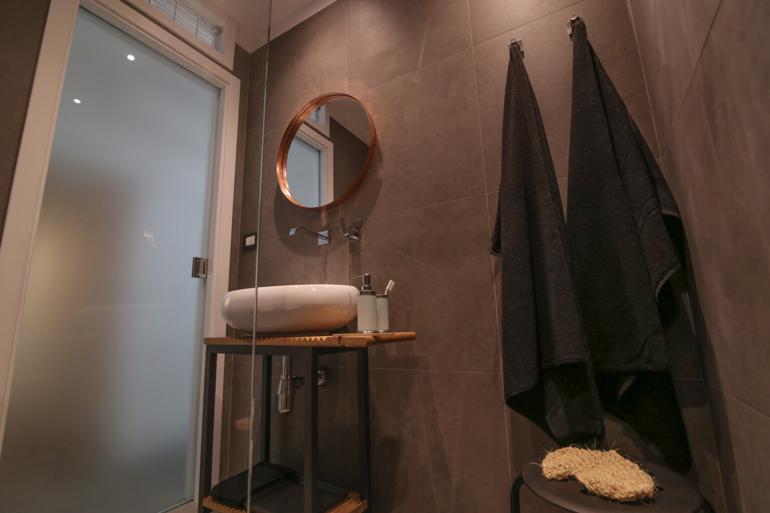 Modern bathroom interior featuring a round mirror, wooden shelf with toiletries, and black towels hanging on hooks against a textured wall. Soft lighting enhances the contemporary design. | Only on SopranoVillas