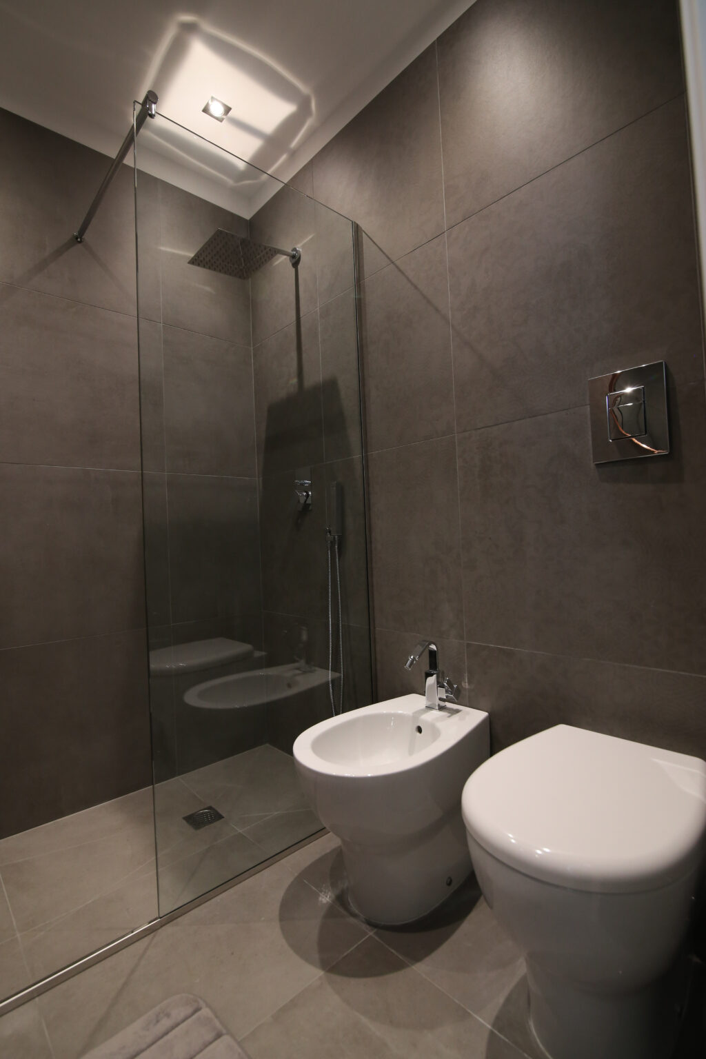 Modern bathroom interior featuring dark tiled walls, a glass shower enclosure, a sleek sink, and a bidet, showcasing a minimalist design. | Only on SopranoVillas