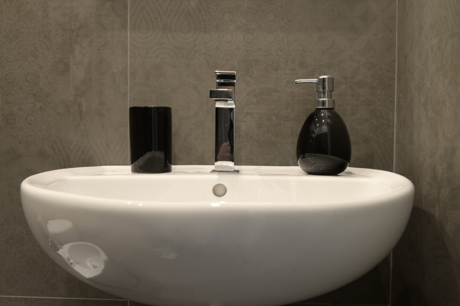 Modern bathroom sink with sleek faucet, black soap dispenser, and minimalist design against a gray tiled wall. | Only on SopranoVillas