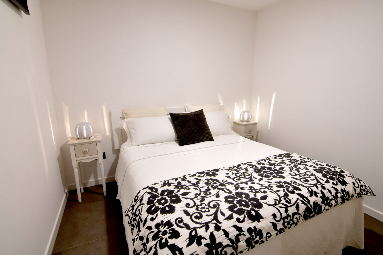 Cozy bedroom featuring a double bed with a decorative black and white throw blanket, bedside tables with lamps, and a minimalist white wall design. Ideal for a comfortable and stylish living space. | Only on SopranoVillas