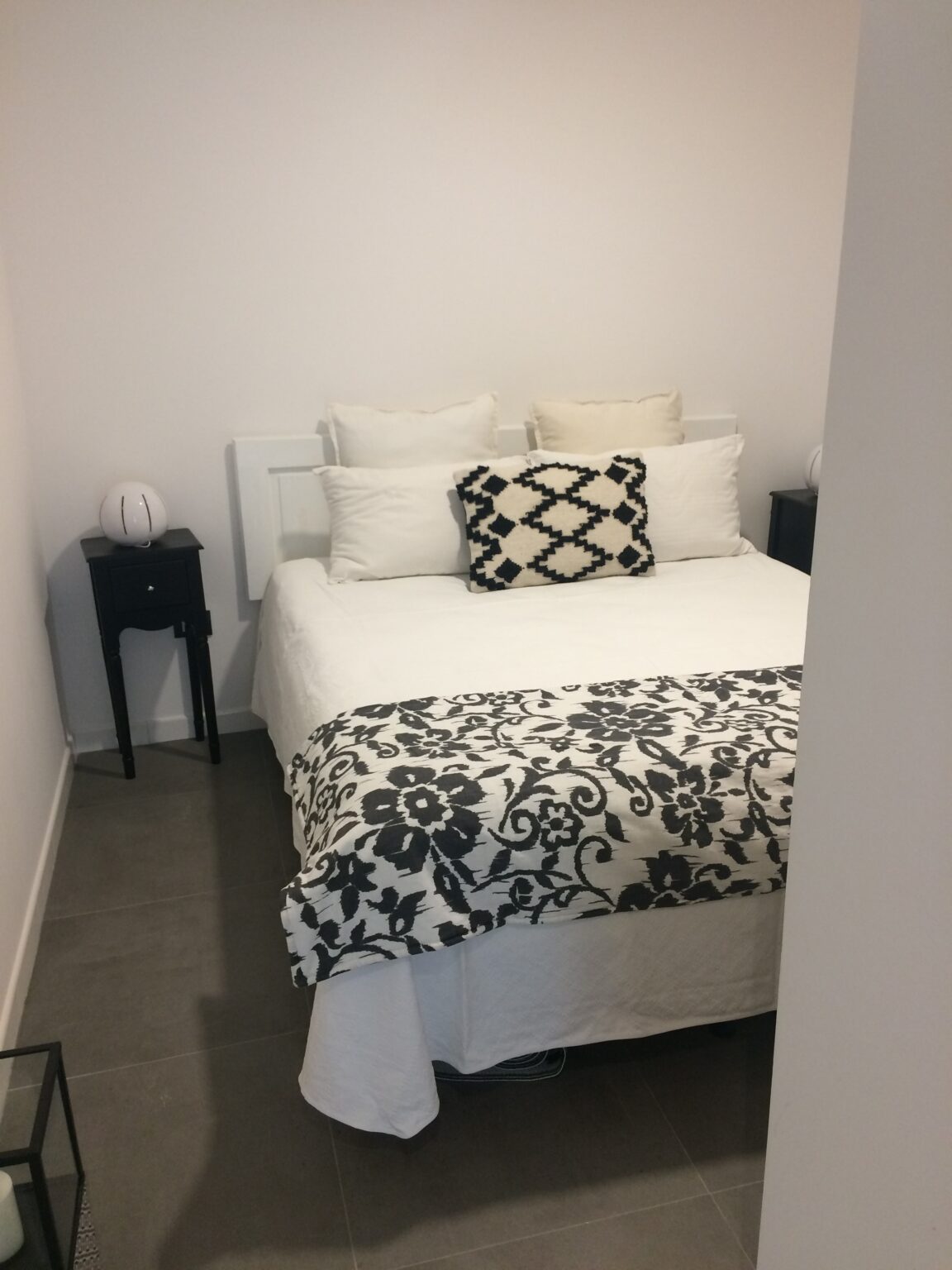 Cozy bedroom featuring a neatly made bed with white linens and a decorative black and white throw blanket, accompanied by stylish pillows. A small black nightstand with a decorative lamp adds to the modern aesthetic. | Only on SopranoVillas