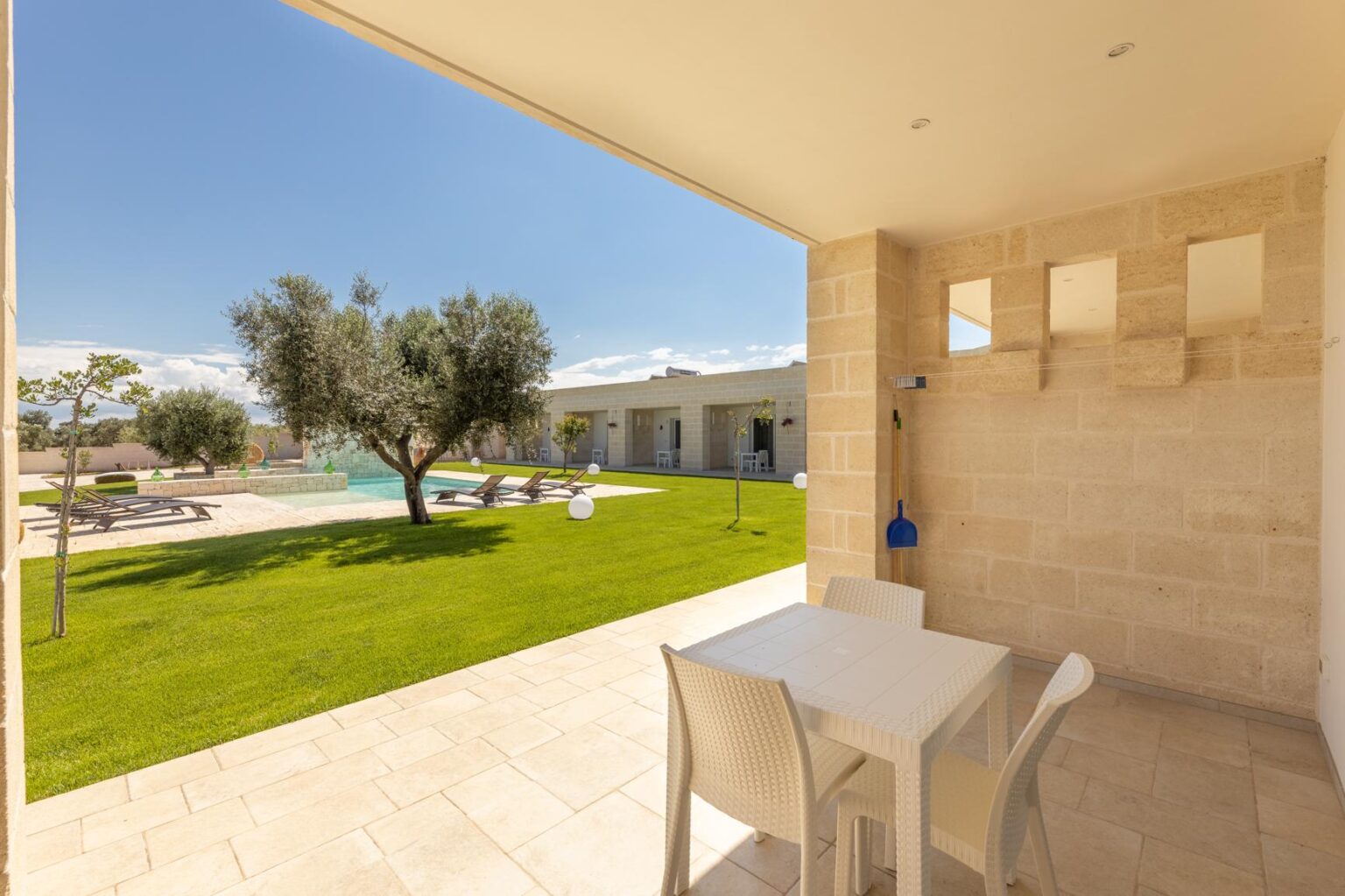 Modern outdoor patio view with a table and chairs overlooking a lush green lawn and swimming pool, surrounded by olive trees and a sunny blue sky. Ideal setting for relaxation and outdoor dining. | Only on SopranoVillas