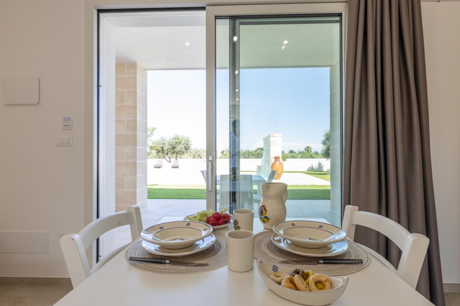 Bright dining area featuring a table set for two, with dishes and a vase, overlooking a scenic outdoor view through large sliding glass doors. | Only on SopranoVillas