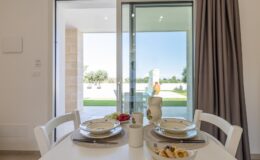 Bright dining area featuring a table set for two, with dishes and a vase, overlooking a scenic outdoor view through large sliding glass doors.