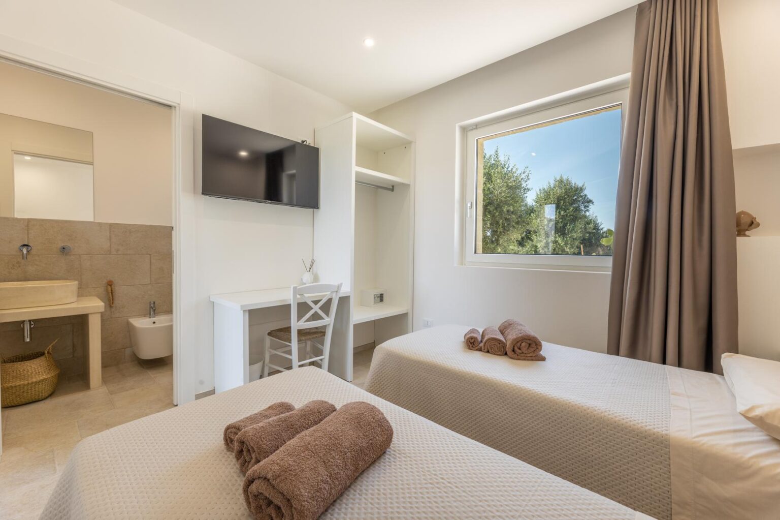 Modern guest room with two twin beds, plush towels, a flat-screen TV, and a window offering a view of greenery, featuring an en-suite bathroom with neutral-toned decor. | Only on SopranoVillas