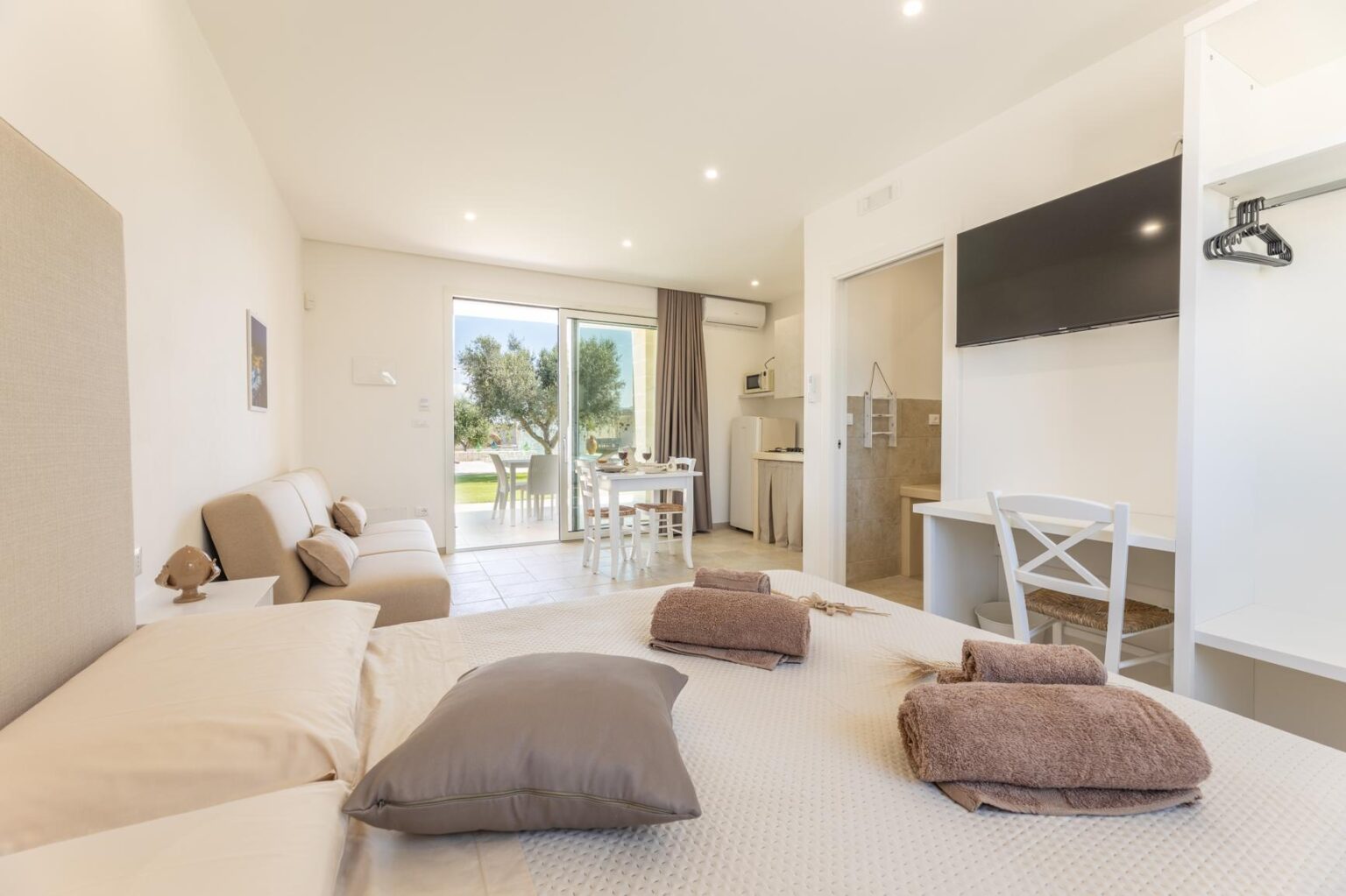 Modern hotel room featuring a cozy bed with decorative pillows, a seating area, and a kitchenette. Large windows provide natural light and a view of the outdoor space, complemented by contemporary furnishings and a flat-screen TV. Ideal for relaxation and comfort during a stay. | Only on SopranoVillas