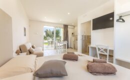 Modern hotel room featuring a cozy bed with decorative pillows, a seating area, and a kitchenette. Large windows provide natural light and a view of the outdoor space, complemented by contemporary furnishings and a flat-screen TV. Ideal for relaxation and comfort during a stay.
