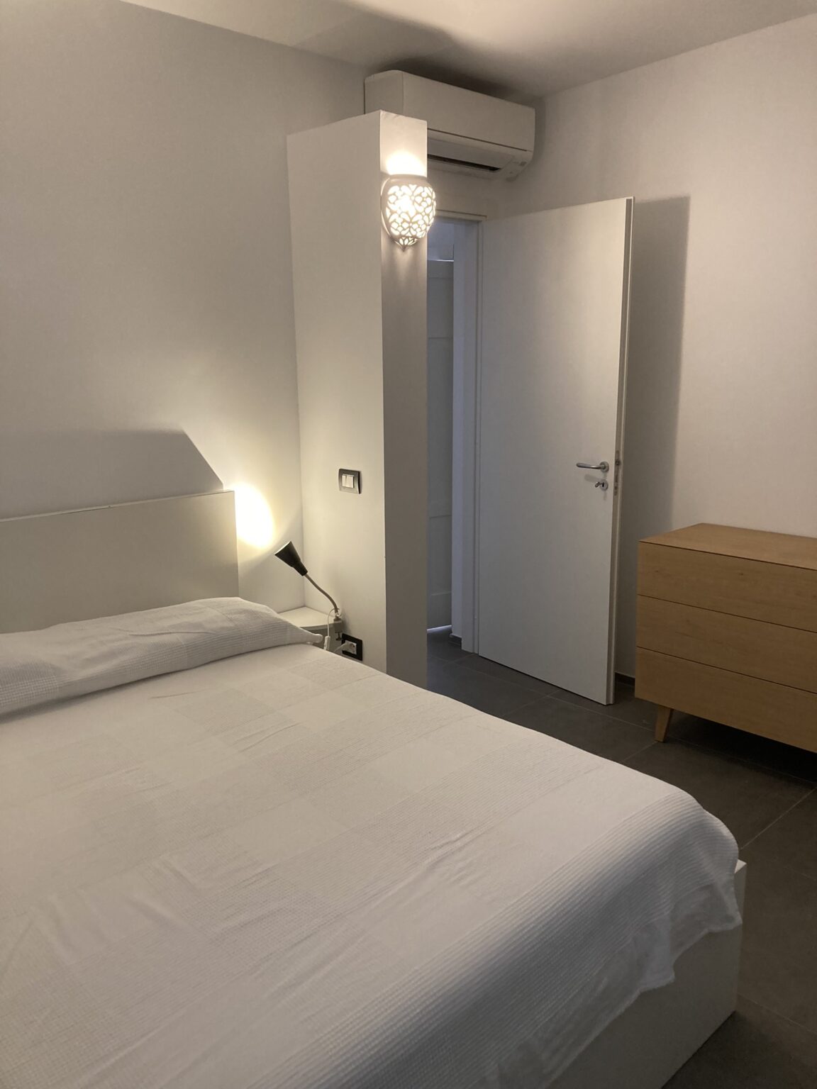 Modern bedroom interior featuring a neatly made bed, a bedside lamp, and a stylish wooden dresser. A door leads to another room, enhancing the spacious feel of the design. Soft lighting creates a cozy atmosphere. | Only on SopranoVillas