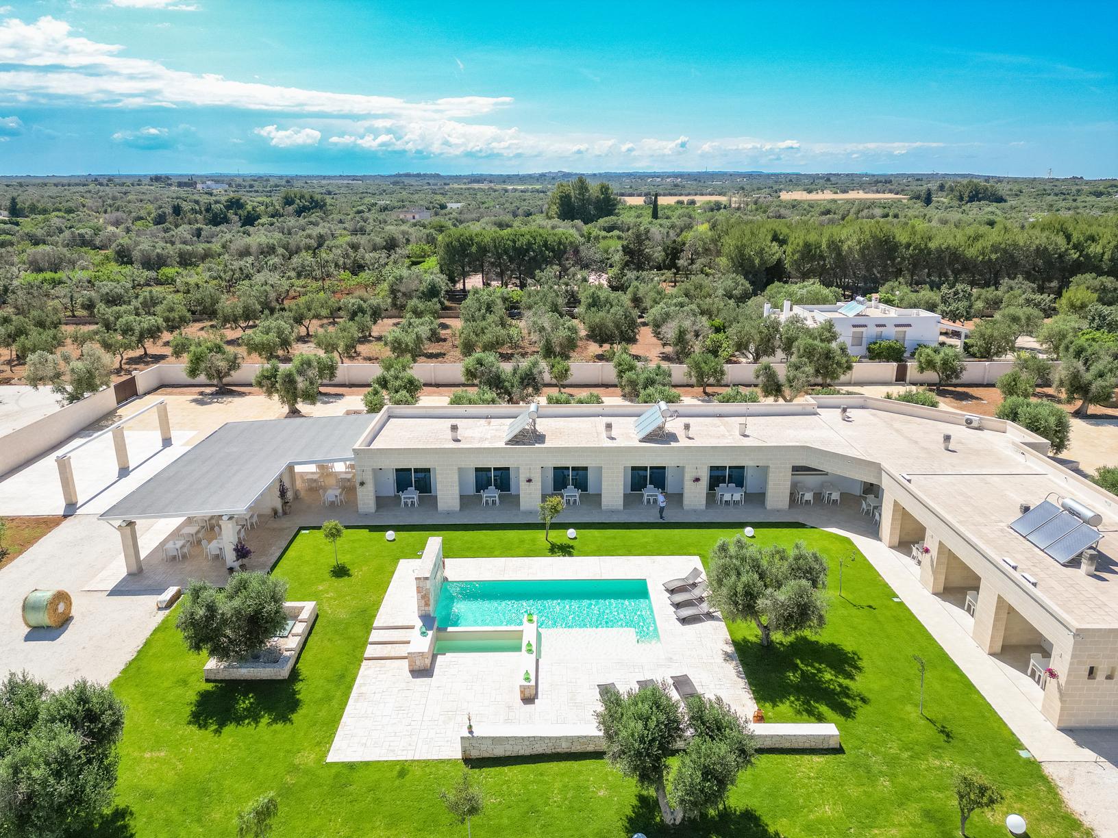 Aerial view of a luxurious villa with a swimming pool, surrounded by lush greenery and olive trees, set against a clear blue sky. The modern architecture features spacious outdoor areas and well-maintained gardens. | Only on SopranoVillas